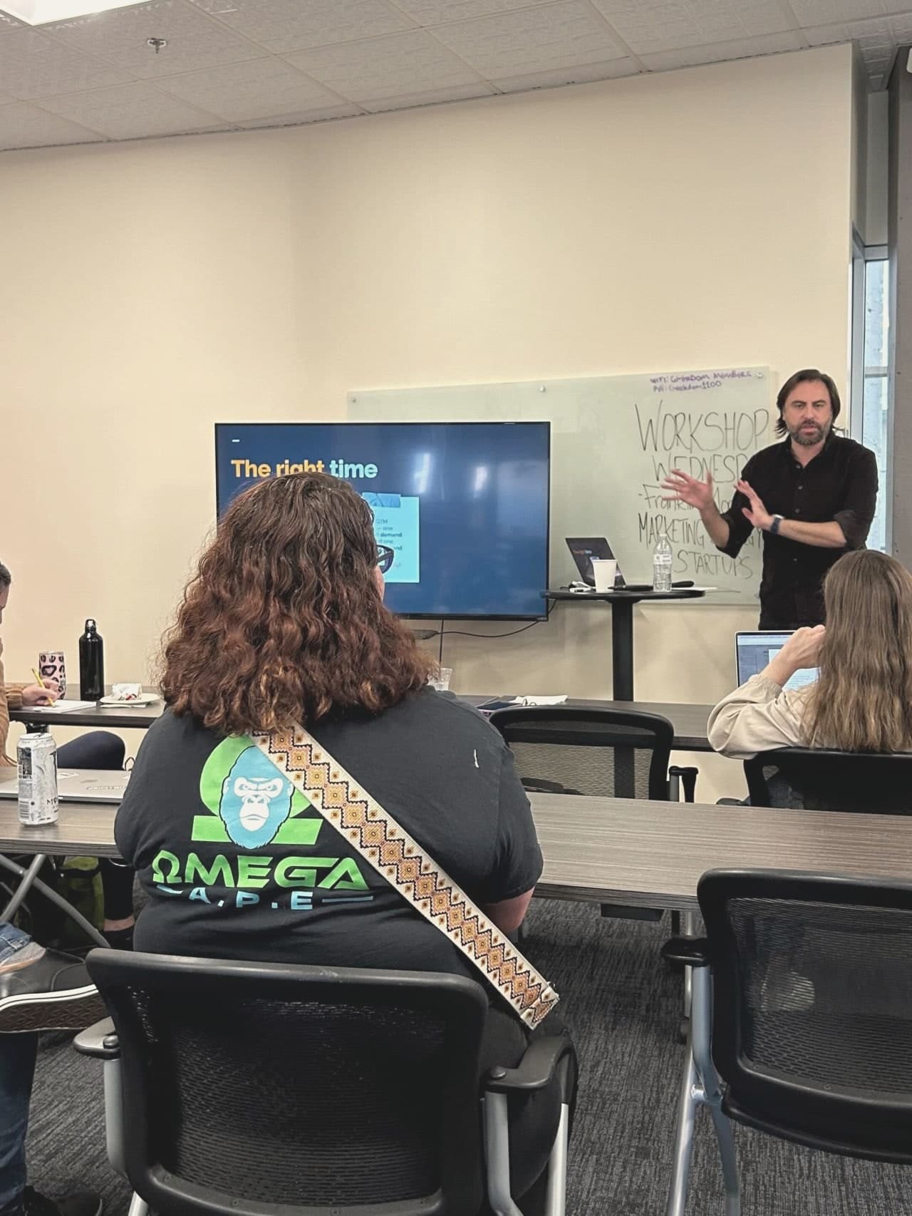 Franklin Morris mid-sentence at a Geekdom workshop, a whiteboard behind him reading Workshop Wednesday, marketing strategy for startups, with founders seated at tables.