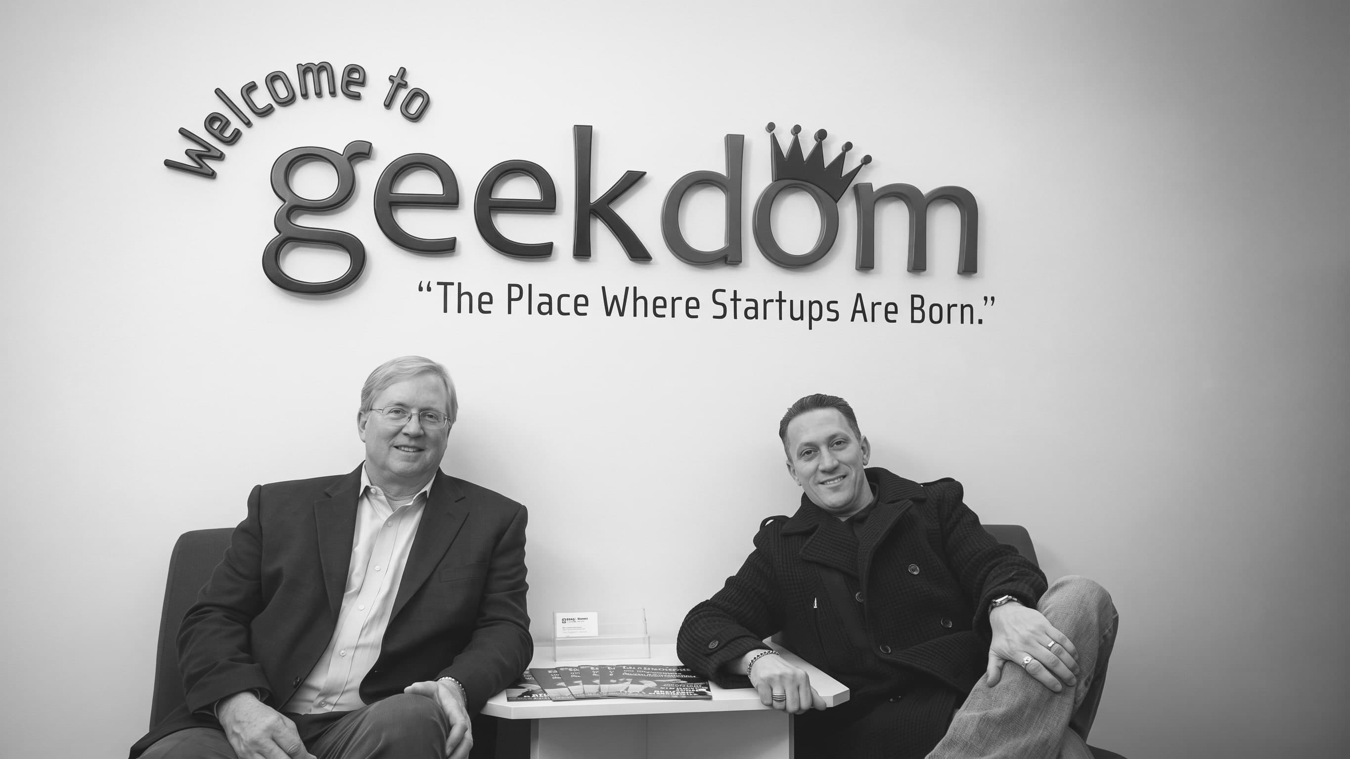 Graham Weston and Nick Longo sitting together in 2011 beneath Geekdom's original wall sign, which reads "Welcome to geekdom — The Place Where Startups Are Born."